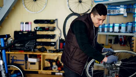 handsome mechanic is cleaning bicycle wheel outside and inside, hub and spokes with piece of cloth and listening to music with earphones. profession and people concept.