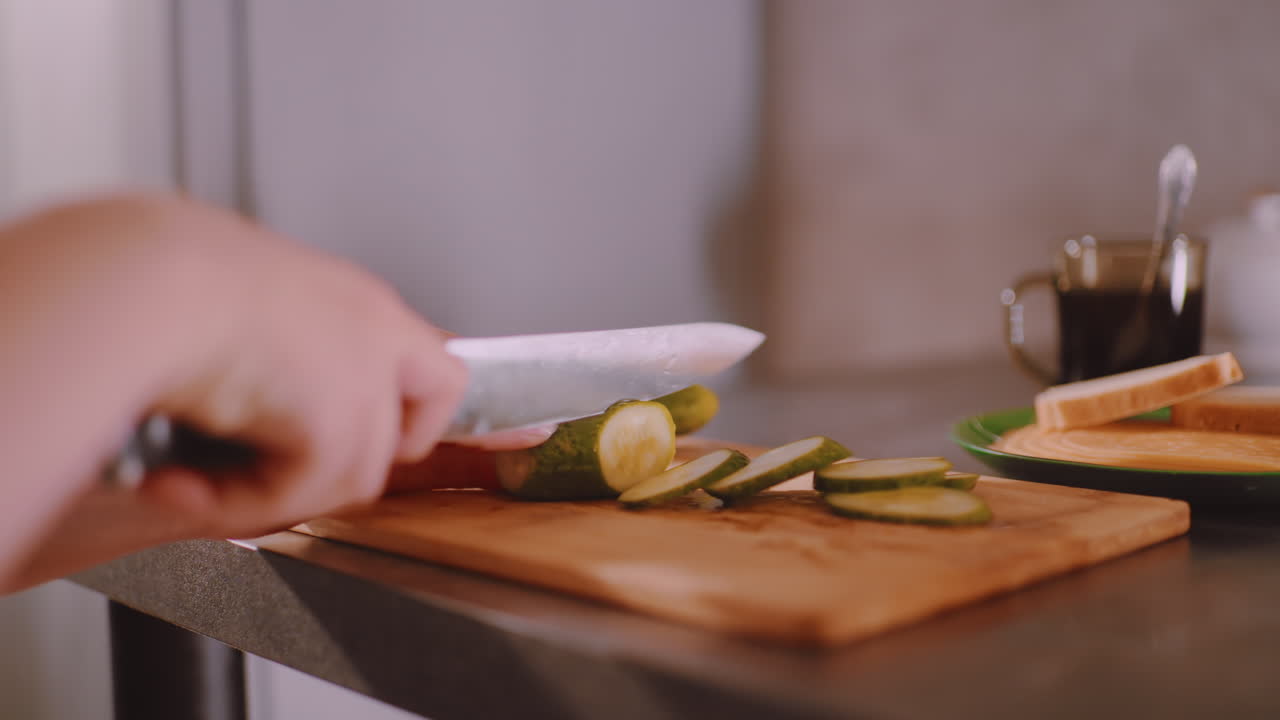 Hand slicing cucumber on wooden cutting board with sharp knife, fresh slices neatly arranged while bread and hot coffee cup sit ahead on table, capturing breakfast preparation in kitchen