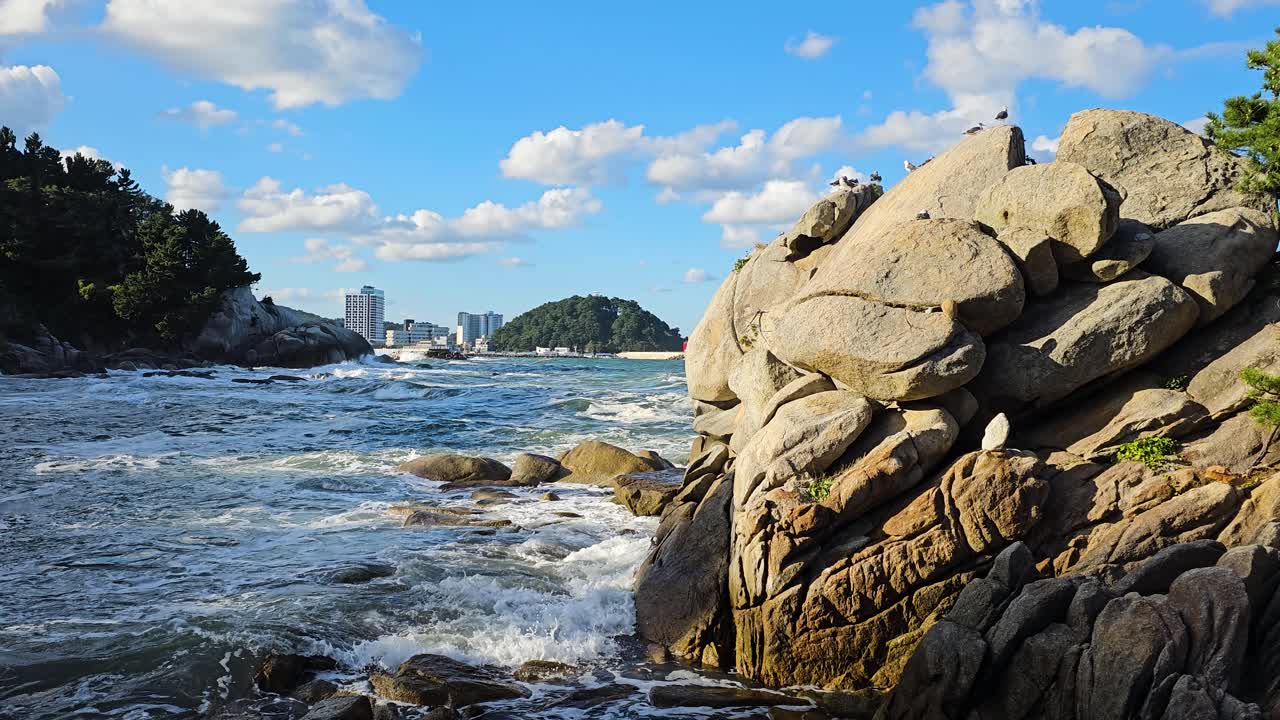 View of the rugged coastal cliffs near Hyuhyuam Hermitage, South Korea, showcasing ocean waves and scenic coastline with buildings in distance on horizon on sunny day