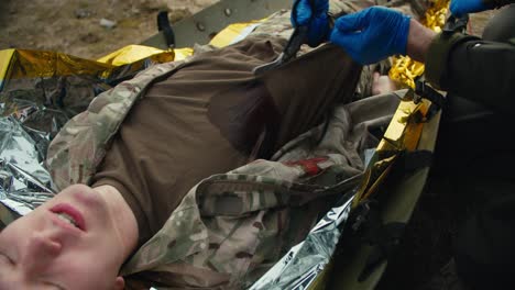 Confident-male-military-man-in-a-dark-green-uniform-and-with-blue-medical-gloves-cuts-the-T-shirt-of-a-wounded-soldier-to-stop-bleeding-during-training-and-combat-exercises-providing-medical-care