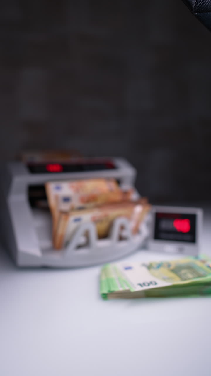 Person places orange bills into cash counting device. Male hand takes counted euro and places on the desk near one hundred stack. Close up. Blurred image. Vertical video.