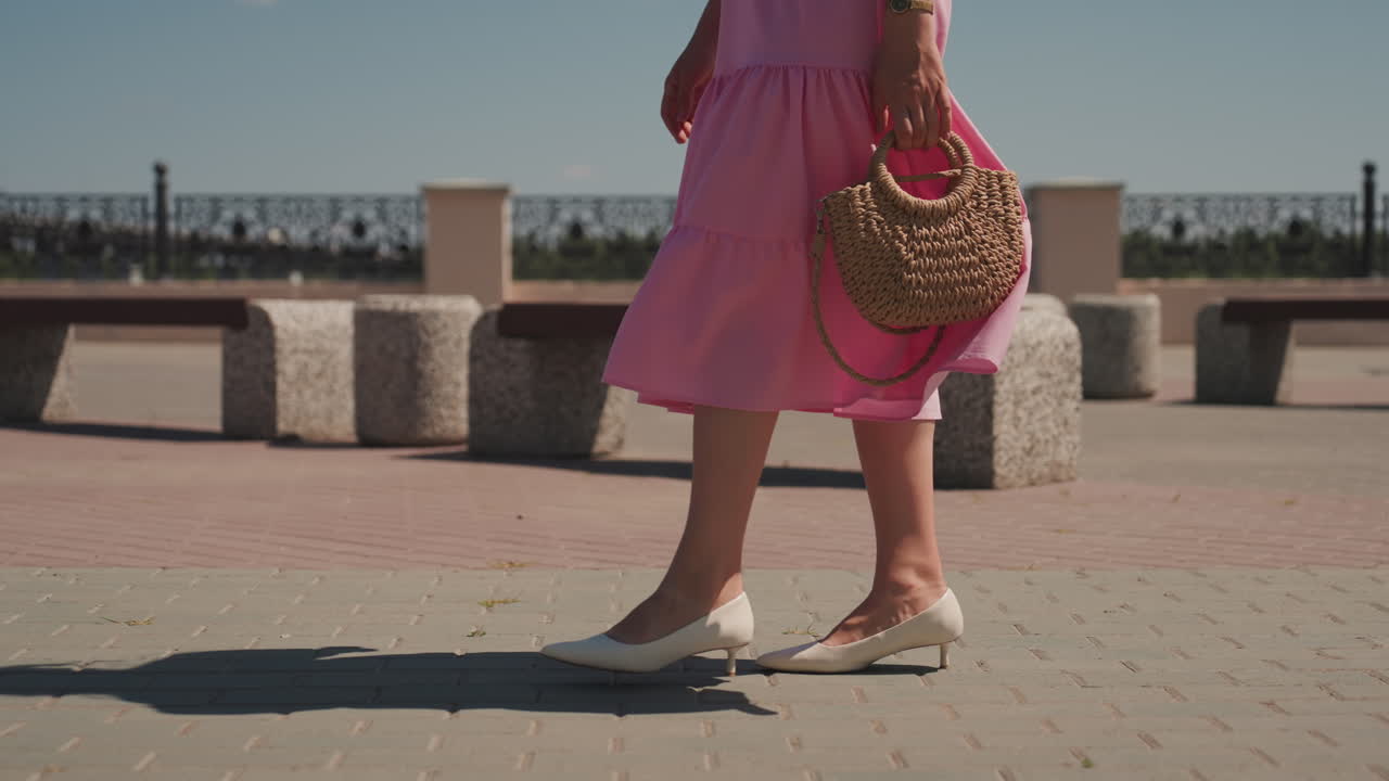 Donna con abito rosa che cammina sul lungomare, primo piano delle gambe e della borsa di paglia, tacchi bianchi, panchine in pietra e fioriere, calda luce solare che proietta lunghe ombre, atmosfera estiva rilassata e passo lento ed elegante