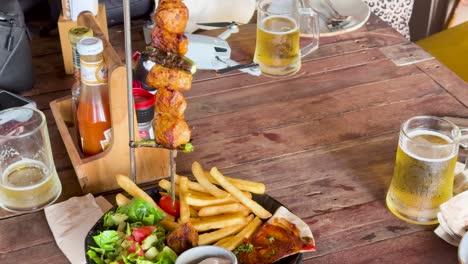 A vibrant meal with shish kebab, fries, salad, and beer on a rustic table at a beach club