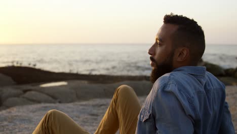 hombre de raza mixta pensativo sentado en una roca en la playa durante la puesta de sol 4k