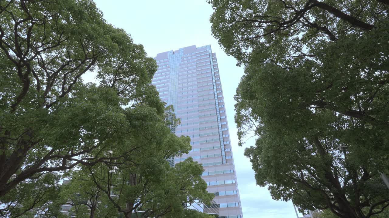 A low-angle shot of a tall, modern skyscraper framed by green trees against an overcast sky