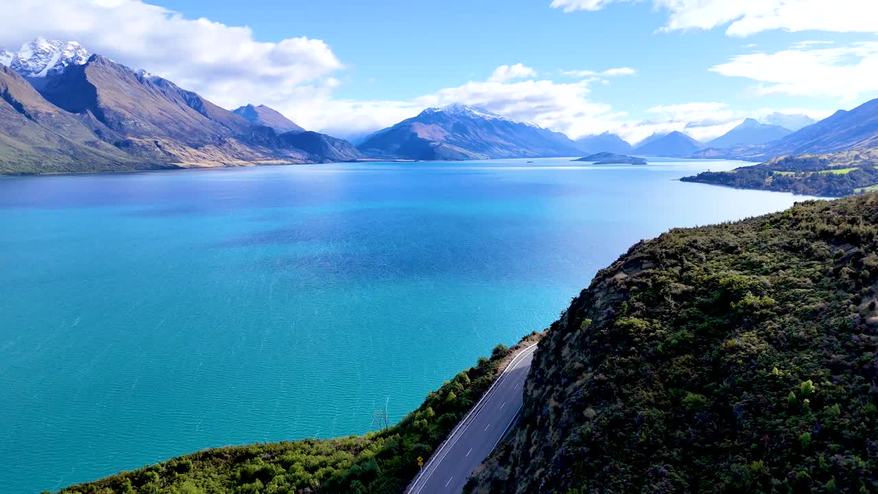 Drone glides above winding road, turquoise lake, and mountains under bright daylight in Queenstown
