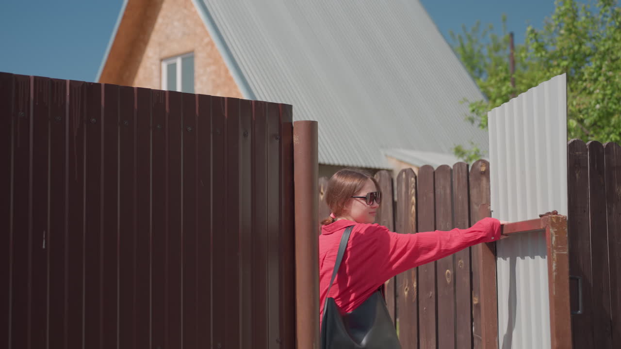 Young woman with long straight hair walks through open gate into residential area as friend walks ahead, creating casual moment of entry under sunny blue sky with house and fence in background
