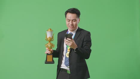 슈트와 타이를 입은 아시아 사업가, 금메달을 들고 금 트로피를 들고 스튜디오에서 녹색 화면 배경에 스마트폰을 사용합니다.