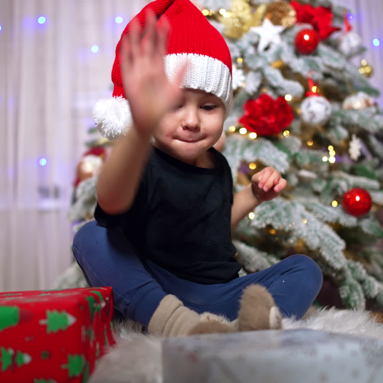 Adorable kid in Santa cap claps his hand by the gift boxes. Baby boy sits on the floor near the Christmas tree.