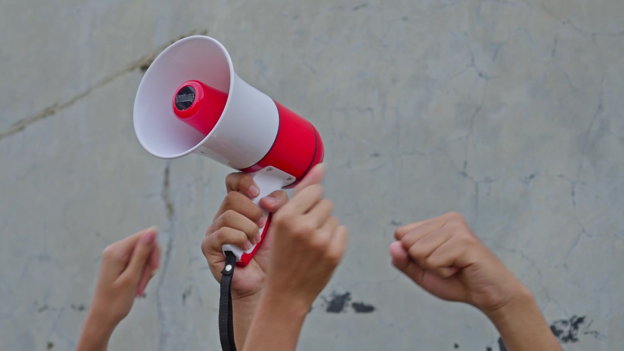 Close-up de mão erguendo megafone cercada por punhos cerrados durante protesto público.
