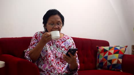 Elderly-asian-woman-sitting-on-the-sofa-while-reading-news-from-her-cellular-phone-and-drinking-coffee-at-home