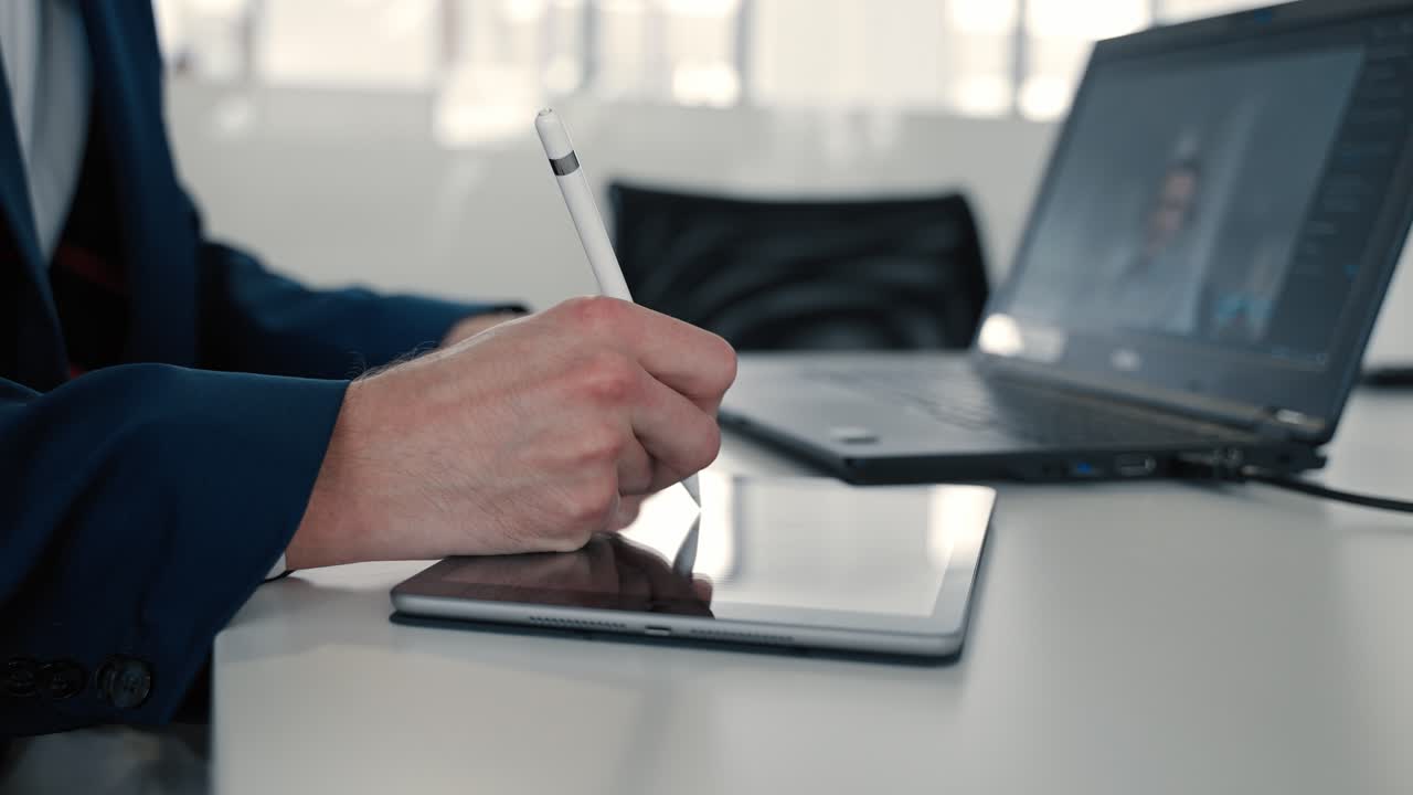 Primo piano della mano di un uomo che prende appunti su un tablet con una penna bianca mentre è in videochiamata sul notebook in un ufficio, indossando un abito blu scuro