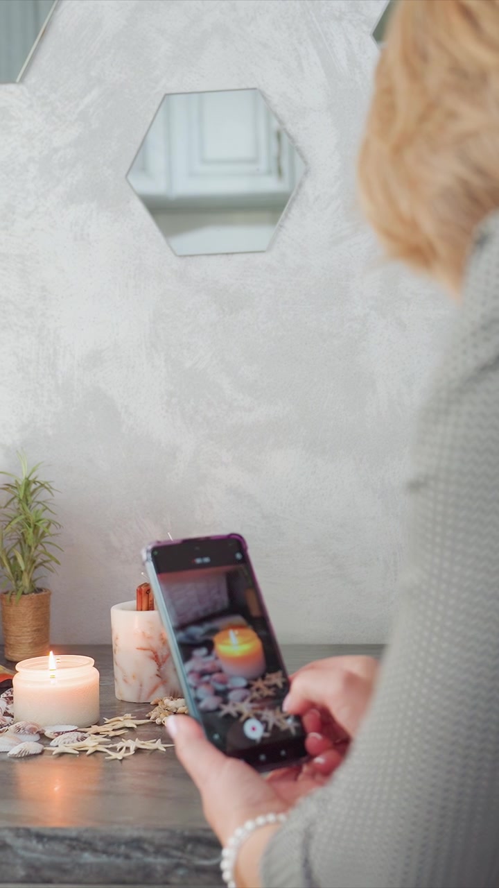 Nærbilde av kvinne som tar bilder med smarttelefonen sin, med fokus på dekorative gjenstander som tente lys, skjell og pent arrangerte trepinner. Scenen har myk belysning mot en teksturert bakgrunn.