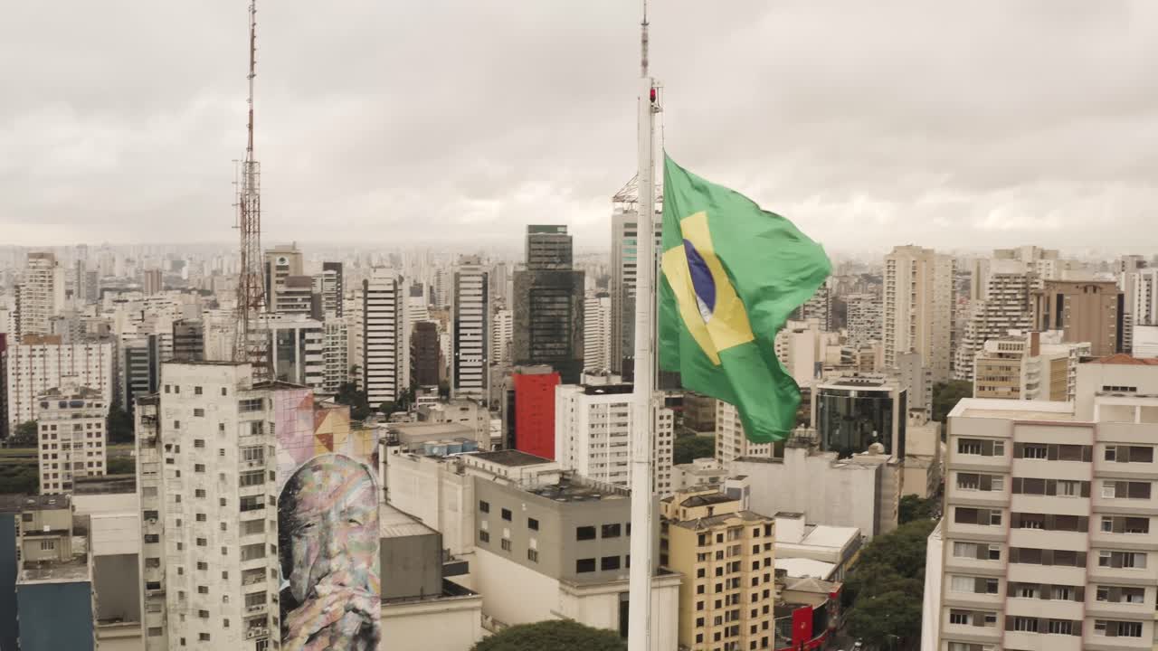 Luftoptagelse i kredsløb af det vajende brasilianske flag på flagstang foran skylinen i Sao Paulo by på en overskyet dag