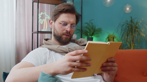 Young-man-relaxing-interesting-book-turning-pages-smiling-enjoying-literature-taking-a-rest-on-sofa