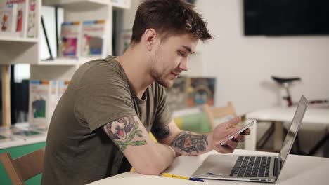 Attractive-programmer-with-tattooes-on-his-arms-scrolling-something-on-his-smartphone-while-sitting-in-a-classroom-beside-laptop.