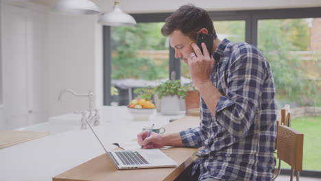 Hombre-Trabajando-Desde-Casa-Usando-Una-Computadora-Portátil-En-El-Mostrador-De-La-Cocina-Hablando-Por-Teléfono-Móvil,-Filmado-En-Cámara-Lenta