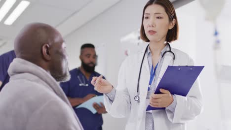 Video-of-diverse-male-patient-and-female-doctor-with-clipboard-talking-in-busy-hospital-corridor