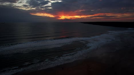 Aerial-flying-over-people-and-surfers-in-ocean-towards-red-sunset