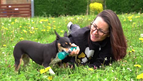 Lady-laying-down-in-grass-playing-with-rescue-dog-and-squeaky-toy,-long-haired-brunette