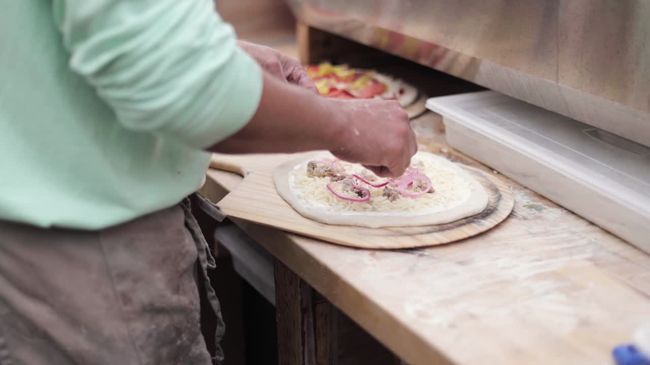 Jovem fazendo pizza em uma pá de madeira, polvilhando vegetais, queijo e carne ao ar livre 1080p 60fps