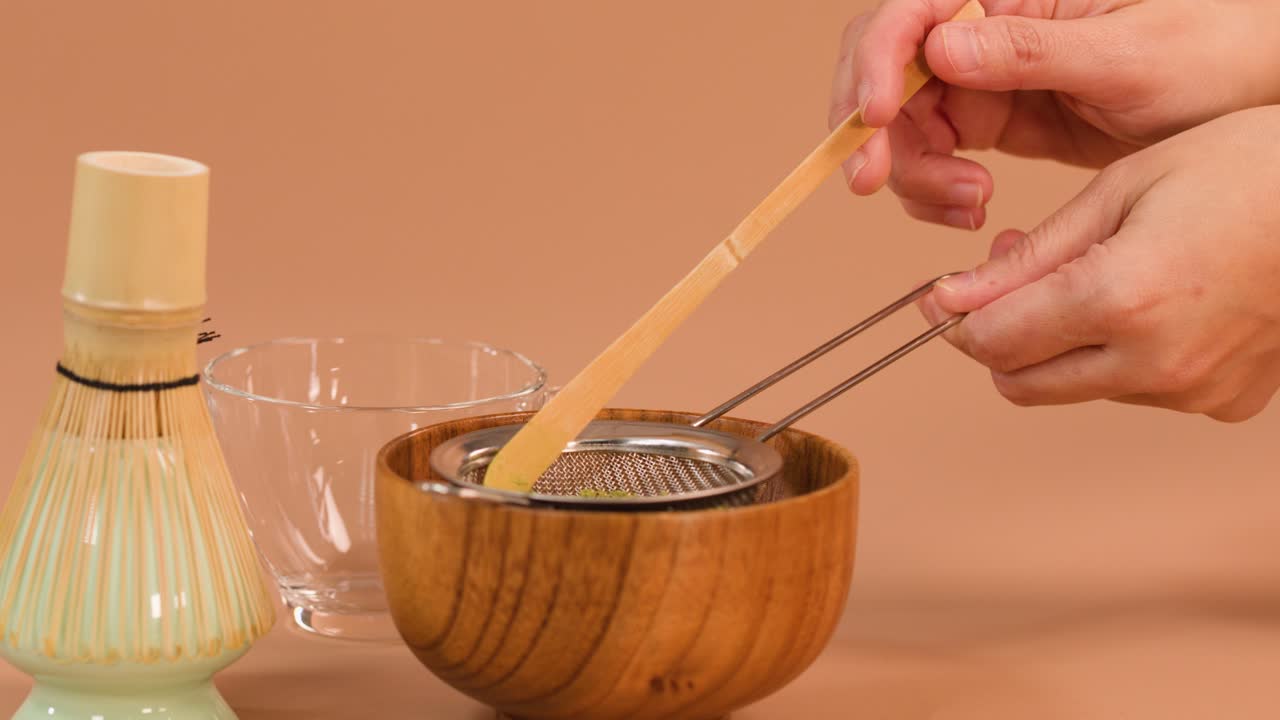 Hand sifts matcha powder into wooden bowl using bamboo scoop and strainer, soft studio lighting