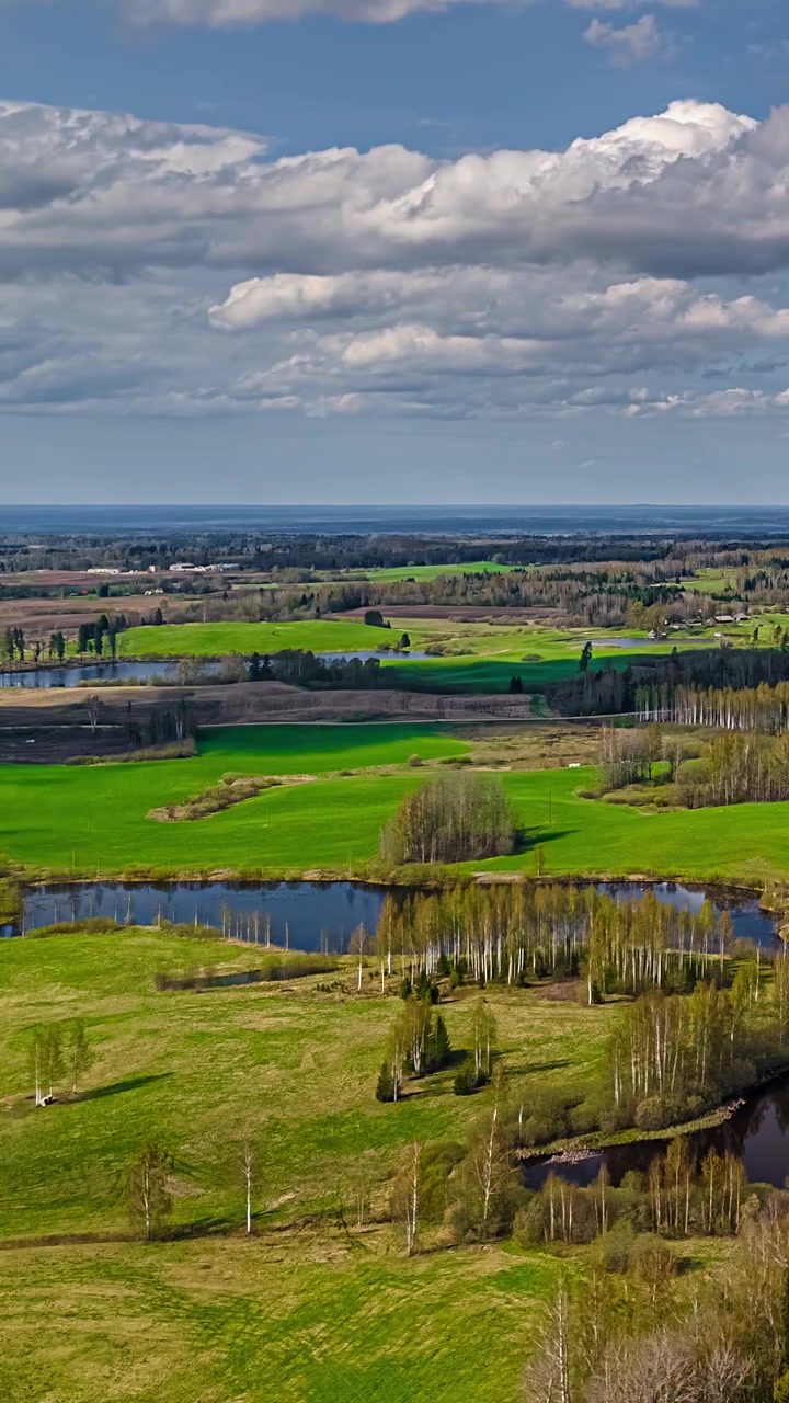 Verticale luchttimelapse boven het Letse platteland terwijl wolken door de lucht drijven, veranderend licht over groene velden, vijvers en met bomen omzoomd landbouwgrond