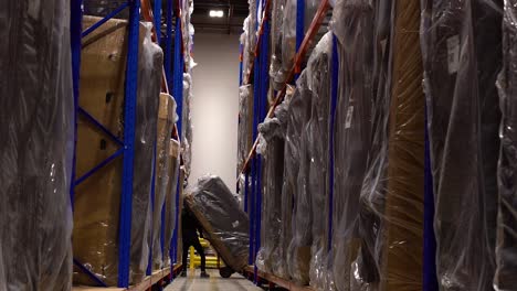 A-Middle-Aged-Warehouse-Worker-Employee-Uses-a-Hand-Dolly-to-Lift-a-Heavy-Cardboard-Industrial-Wrapped-Inventory-Furniture-Package-Out-of-Blue-and-Orange-Distribution-Center-Racking