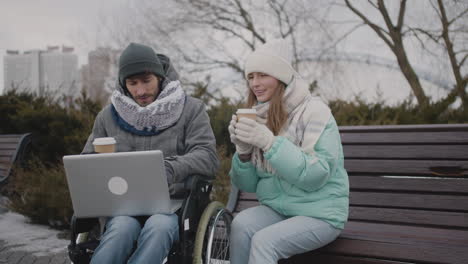 disabled man in wheelchair watching something funny on laptop computer with his friend at urban park in winter