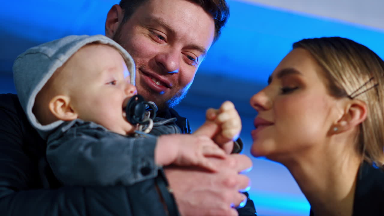 Little baby in a hood with pacifier in mouth held by dad. Loving mm leans to kiss her son. Low angle view.