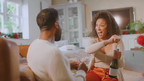 loving couple celebrating opening bottle of champagne around christmas tree at home
