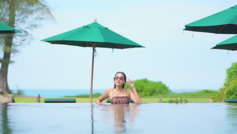 Modelo-Asiático-De-Pie-En-La-Piscina-Con-Gafas-De-Sol-Mirando-El-Paisaje-Del-Paraíso-Tropical-Del-Complejo-Isleño-Con-Sombrillas-Verdes-Y-Oceánicas-En-El-Fondo