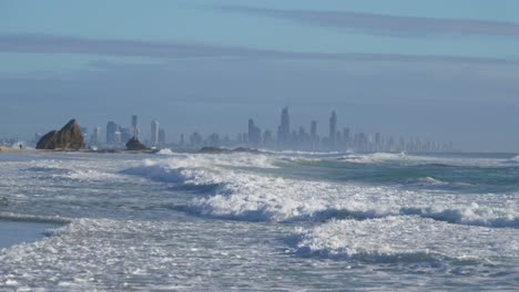 Surfwellen,-Die-An-Einem-Sommermorgen-über-Das-Meer-Am-Strand-Von-Currumbin-Rollen---Skyline-Der-Goldküste-Im-Nebligen-Hintergrund---Weitschuss