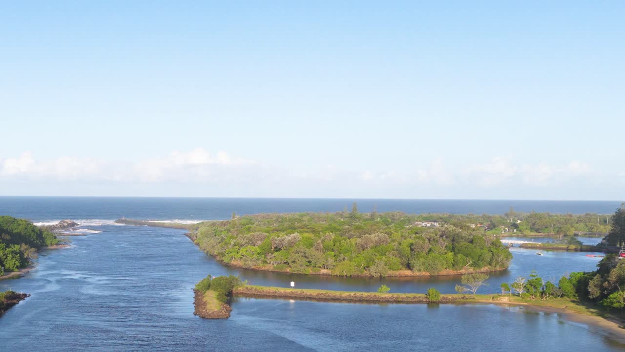 Aerial footage captures a serene river and lush island under clear skies at Brunswick Heads, NSW, Australia