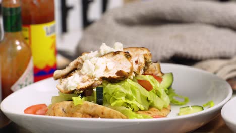 primer plano de una ensalada de pollo a la parrilla en un recipiente poco profundo mientras las manos enguantadas de un cocinero desmenuzan el queso feta en un recipiente poco profundo