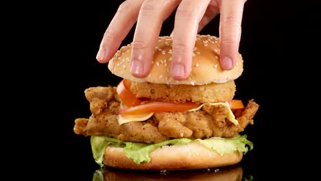A hand lifts a sesame bun chicken burger with lettuce, tomato, cheese, and crispy onion ring against a black background, using smooth overhead lighting and steady camera framing