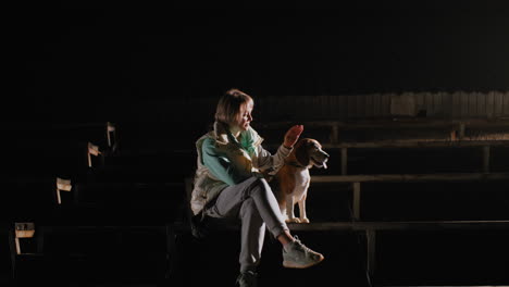canine specialist seated on staircase at night touching ears of beagle dog playfully as bright spotlight illuminates surroundings creating calm bonding moment under empty dark stadium setting