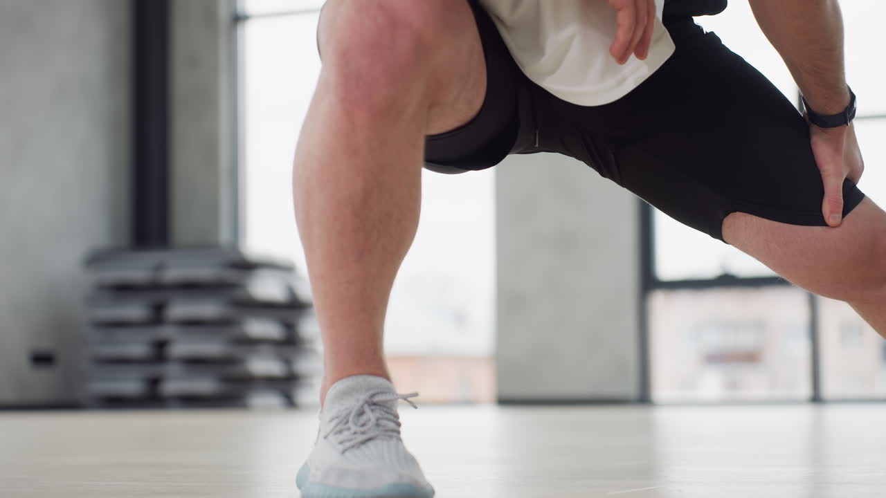 Student stretching left leg in bright fitness studio wearing white tank and black shorts performing side lunge warm up on light wooden floor under large windows with hanging straps