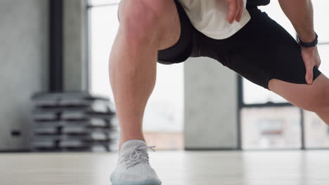 student stretching left leg in bright fitness studio wearing white tank and black shorts performing side lunge warm up on light wooden floor under large windows with hanging straps