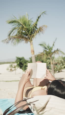 vertical video of biracial woman reading book lying on beach in sun, slow motion