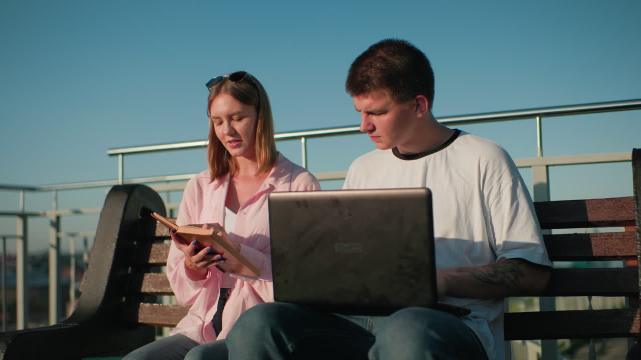 estudiantes de secundaria sentados en un banco al aire libre estudiando, el hombre se centró en escribir en la computadora portátil con la mano tatuada mientras miraba lo que la mujer está mostrando del libro
