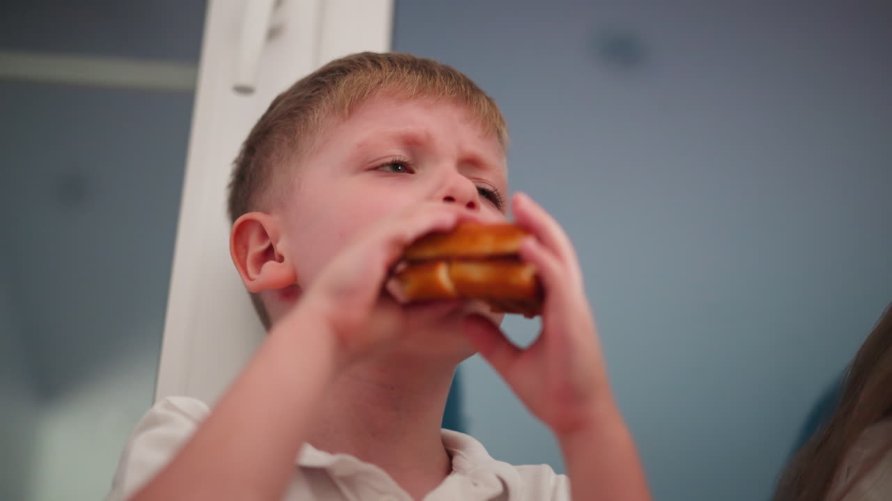 Close-up of young boy holding dessert with both hands as he takes a big bite, partially showing sister beside him with soft blur background of indoor glass door and evening lighting ambiance