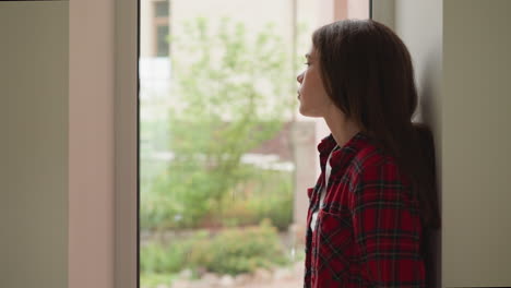 una mujer joven está de pie junto a una ventana, mirando hacia afuera y pareciendo pensativa.