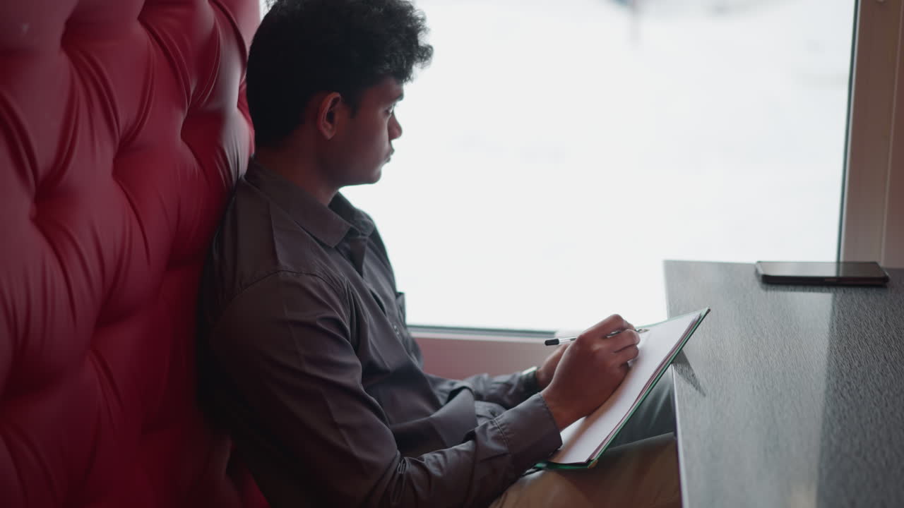 Student seated beside window drawing in sketchbook, holding pen with focused expression, soft natural light from snowy outdoors creates peaceful indoor study environment