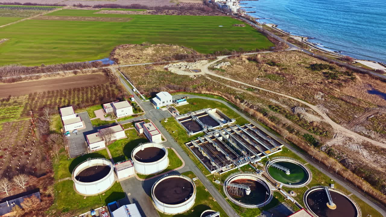 Water treatment facility located near the coast with circular tanks and green areas containing roads and buildings, taken during daylight hours