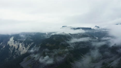 Panoramic-aerial-overview-of-wispy-clouds-covering-mountain-ridges-and-valleys-with-exposed-rocky-slopes-between-forest-pockets