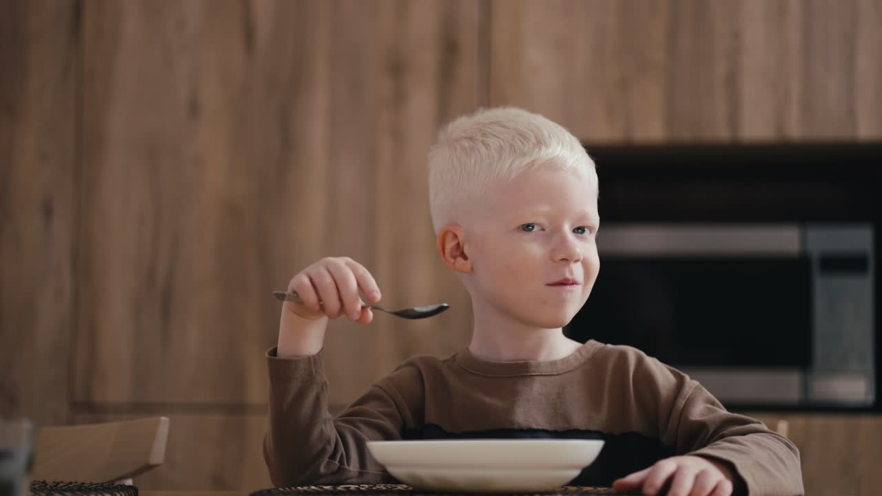 Portrait of a happy albino boy with white hair who has breakfast in the kitchen and eats oatmeal with a spoon while sitting at a modern table in a modern kitchen at home