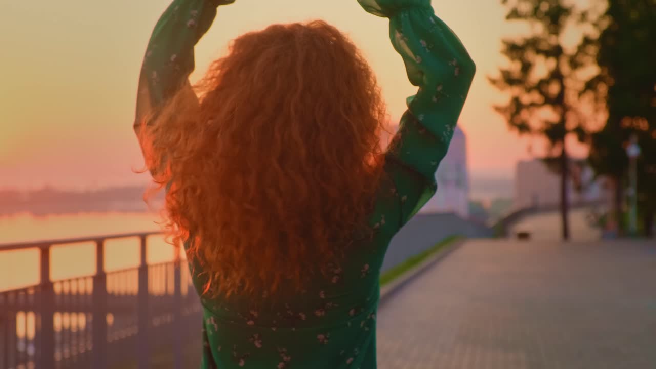 A Golden Sunset: A Woman with Curly Red Hair Enjoys the Serenity of Dusk While Strolling Along a Scenic Path in a Tranquil Urban Landscape