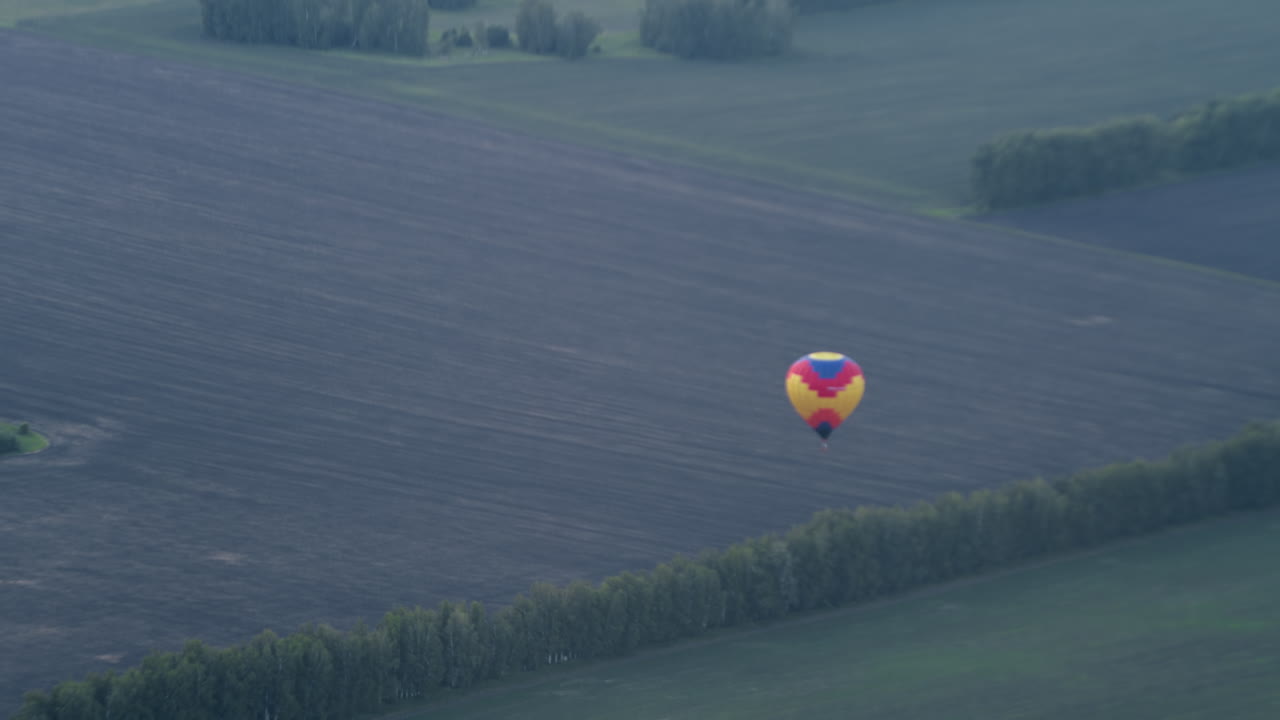 Distant view of colourful hot air balloon descending over vast tilled field bordered by tree lines, wicker basket swinging beneath rippled canopy as gentle breeze guides tranquil aerial descent