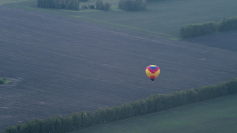 distant view of colourful hot air balloon descending over vast tilled field bordered by tree lines, wicker basket swinging beneath rippled canopy as gentle breeze guides tranquil aerial descent